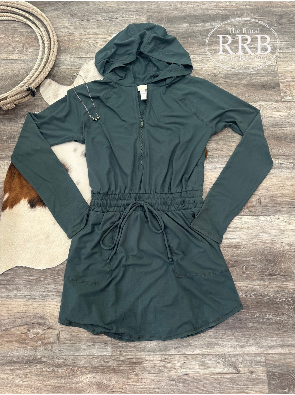 Step out in style with The Hannah Long Sleeve Quarter Zip Hooded Romper Dress in Blue Spruce. Crafted from buttery soft, stretchy fabric, this romper features a smocked elastic drawstring waist and hidden shorts with a 4-inch inseam for added comfort. The stylish design includes rectangle stitched side pockets and a rounded hem, making it perfect for any occasion. Available in sizes Small (2-4), Medium (6-8), and Large (9-11). Shown with The Everyday Navajo necklace.