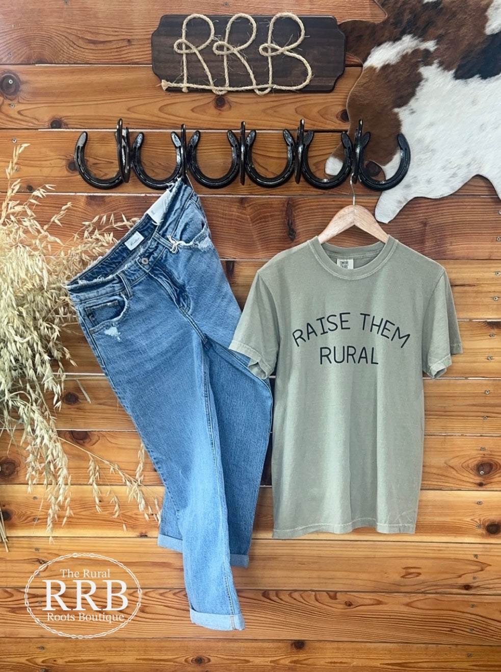 Embrace rural pride with the Raise Them Rural T-shirt in khaki green featuring bold black text. Crafted from 100% premium cotton blend for ultimate comfort, this unisex-fitting shirt is available in Small (3-5), Medium (6-8), Large (9-11), X-Large (12-15), and XXL (16-18). Shown paired with the Decatur High Rise Wide Leg Jean for a stylish look.