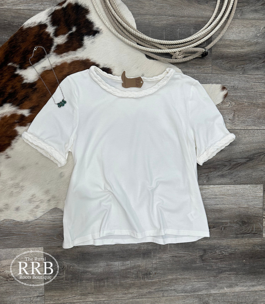 Discover The Chappell Short Sleeve Top in White, featuring a stylish braided knot trim on the neckline and sleeves. Made from a comfortable blend of 95% cotton and 5% spandex, this top offers a flattering fit that's perfect for summer outings. Available in small, medium, and large, it's a versatile addition to your wardrobe, ideal for pairing with shorts, jeans, or skirts.